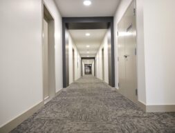 A long, narrow hotel corridor lined with closed doors and carpeted floors, illuminated by overhead lights.