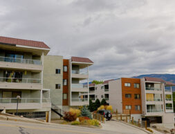 Modern apartment buildings with balconies on a hilly street under a cloudy sky, with distant mountains and a parked car visible.
