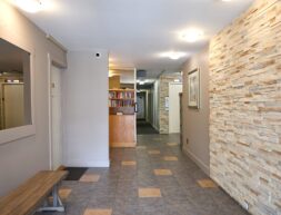 Interior of a building lobby with tiled flooring, a stone wall, a wooden bench, and bookshelves filled with books.