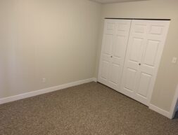 A plain, empty room with beige walls and beige carpet, featuring a closed white double door closet on one wall.